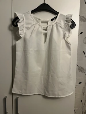 Vit ärmlös blus med volang från SHEIN - Stilren vit blus från SHEIN med volangdetaljer vid axlarna och snygg öppning framtill och baktill. Blusen är ärmlös och har dekorativa knappar bak. Perfekt till jeans eller kjol för en fräsch look.