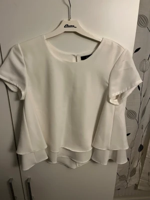 Vit volangblus med dubbla lager - Säljer en stilren vit blus med korta ärmar och rund hals. Blusen har dubbla lager med volangdetalj nedtill som ger ett flowy intryck. Knäpps med en knapp i nacken. Perfekt att styla till jeans eller kjol.
