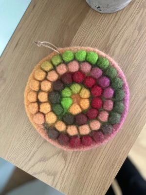 Rund färgglad ullunderlägg - Unikt runt underlägg i tovad ull med färgglada bollar i orange, grönt, rosa, rött och brunt. Underlägget har ett mönster i cirklar och en liten ögla för upphängning. Perfekt som dekoration eller för att ställa varma saker på bordet.