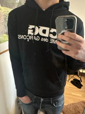 Svart CDG hoodie med vit logga - Snygg svart hoodie från Comme des Garçons med stor vit CDG-logga på bröstet. Klassisk modell med huva och dragsnören samt en stor magficka. Perfekt för dig som gillar streetwear och vill sticka ut med en ikonisk design.