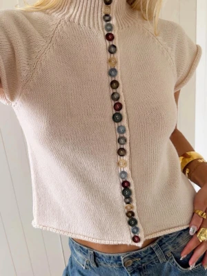 Beige stickad topp LÄS BESKRIVNING  - INTE TILL SALU. Vill bara hitta liknande toppar.!!!!! Trendig beige stickad topp med korta ärmar och hög krage. Framsidan pryds av en rad färgglada knappar i olika nyanser. Perfekt för dig som gillar unika detaljer och en avslappnad men stilren look. Passar snyggt till jeans eller kjol.