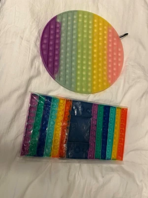 Två stora regnbågsfärgade pop it nypris 400kr - Säljer två stora pop it-leksaker i regnbågsfärger, en rund och en rektangulär. Perfekta för att stressa ner eller bara ha kul med. Tillverkade i mjuk silikon och har tydliga färgskiftningar i lila, blått, grönt, gult, orange och rött.