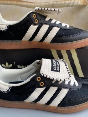 Adidas Wales Bonner svartvita sneakers - Unika Adidas Wales Bonner sneakers i svart och vitt med klassiska tre ränder på sidan, guldfärgade öljetter och detaljerad söm längs kanterna. Ovandelen är i läder och mocka, med gummisula i brun färg. Tungan har Wales Bonner-logga och snörningen är vit med svart mönster.