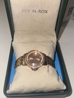 Guldfärgad klocka  - Snygg guldfärgad klocka från Ivy N Rox med metallarmband och rund urtavla i matchande ton. Urtavlan har en stilren design och ramen är dekorerad med små stenar för extra bling. Perfekt accessoar för att lyfta din outfit. Går att förlänga storleken.