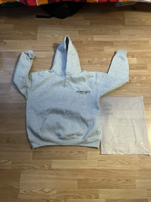 Grå Essentials Fear of God hoodie - Snygg grå hoodie från Essentials Fear of God med svart logga på bröstet och stor text på ryggen. Klassisk känguruficka framtill och stor huva. Perfekt för en avslappnad och trendig streetwear-look.