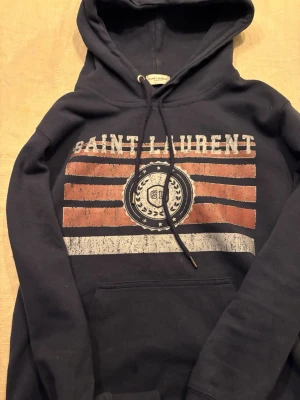 Mörkblå Saint Laurent hoodie - Snygg mörkblå hoodie från Saint Laurent med tryck i vitt och rött framtill. Storlek S,Hoodien har en stor ficka på magen, dragsko i huvan och ribbade muddar. Perfekt för en avslappnad och trendig stil. Materialet känns mjukt och bekvämt.