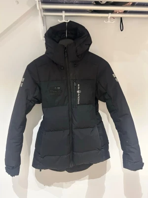 Svart pufferjacka från Sail Racing - Säljer en svart pufferjacka från Sail Racing med hög krage och stor huva. Jackan har logga på bröstet och ärmen, samt dragkedja framtill. Lite indragen vid midjan för en snygg look. Står att det är storlek M men skulle definitivt passa S bättre.