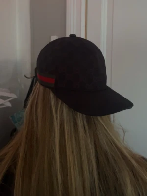 Svart Gucci keps med röd-grön rand - Snygg svart keps från Gucci med klassiskt monogrammönster och en ikonisk röd-grön rand på sidan. Kepsen har böjd skärm och justerbar rem bak. Tillverkad i slitstarkt tyg som ger en exklusiv känsla och passar perfekt till streetwear-stilen.