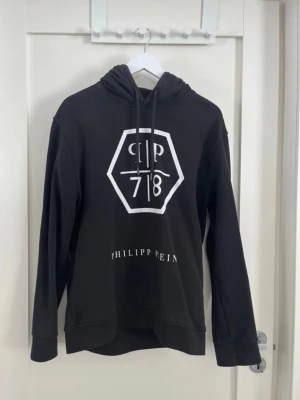 Svart hoodie från Philipp Plein - Svart hoodie från Philipp Plein med stor vit logga och text framtill. Klassisk modell med huva och dragsko. Tillverkad i mjukt material som passar perfekt till en avslappnad stil. Snygg och enkel att matcha med jeans eller joggers.