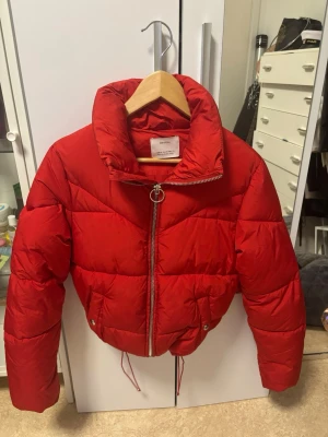 Röd pufferjacka från Bershka - Snygg röd pufferjacka från Bershka med dragkedja med ringdetalj. Jackan har en croppad passform och två fickor med tryckknappar. Man kan croppa den till den längd man vill ha den. Perfekt för kalla vinterdagar och ger en cool streetstyle-look. 