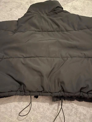 Svart pufferjacka från Divided - Säljer en svart pufferjacka från Divided med hög krage och dragsko i nederkant. Jackan har en klassisk quiltad design och är perfekt för kalla dagar. Materialet är syntetiskt och jackan har en oversized look som är riktigt trendig just nu.