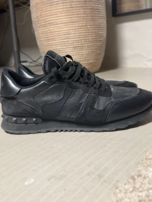 Valentino Garavani Rockrunner sneakers - Säljer ett par svarta Valentino Garavani Rockrunner sneakers med coola nitar på hälen och camouflagemönster i mocka och skinn. Skorna har snörning och en platt sula, perfekt för dig som gillar stilrena och edgy detaljer.