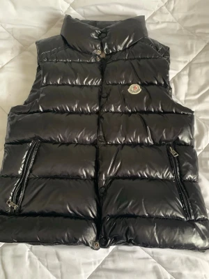 Svart dunväst från Moncler - Snygg svart dunväst från Moncler med glansig finish och hög krage. Västen har två dragkedjefickor framtill och Moncler-logga på bröstet. Perfekt för lager på lager och håller dig varm med sin vadderade design.
