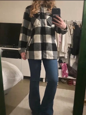 Kappa rutig  - Kappa i svartvitt rutigt mönster med krage och knäppning framtill. Två stora bröstfickor med lock och knapp. Långärmad modell i mjukt, tjockare tyg som ger en avslappnad och trendig look.