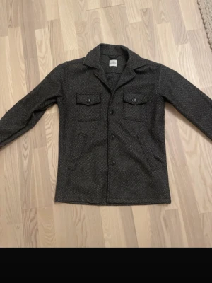 Mörkgrå overshirt med fickor - Stilren mörkgrå overshirt i ullmix med diskret mönster. Två bröstfickor med lock och knappar samt två snedställda sidofickor. Klassisk krage och knäppning framtill. Perfekt lager på lager-plagg för en trendig look.