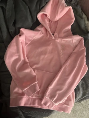 Rosa Juicy Couture hoodie i velour - Säljer en ljusrosa hoodie från Juicy Couture i mjuk velour med huva och magficka. Framsidan har diskret JC-logga i strass och baksidan har Juicy Couture i stora glittriga bokstäver. Perfekt för en chill och trendig look.Tröjan är i ett mycket bra skick. Den är i storlek 14-15 år.
