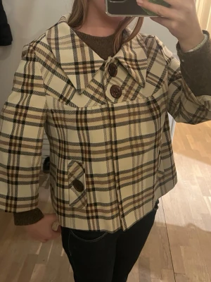 Rutigt cape-inspirerat kappa - Snygg kappa med rutigt mönster i beige, brunt och vitt. Modellen är cape-inspirerad med vida ärmar, stora bruna knappar och bred krage. Perfekt för dig som gillar retrostil och vill sticka ut. Fickor framtill med knappdetaljer. Materialet känns kraftigt och håller värmen.