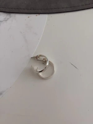 Silvriga hoops örhängen - Säljer ett par stilrena silvriga hoops örhängen i metall. De har en klassisk rund form och blanka ytor som ger en modern vibe. Perfekta för dig som gillar minimalistiska accessoarer och vill ha något som funkar till många olika looks. Aldrig använda, kan tappa färg från Kappahl.