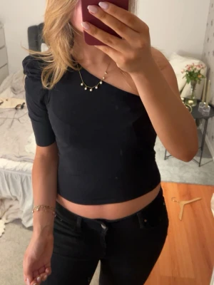 Svart one shoulder croppad topp - Trendig svart croppad topp med one shoulder-design och kort ärm. Snygg passform som framhäver axlarna och passar perfekt till höga jeans eller kjol. Enkel och stilren look som är lätt att matcha.