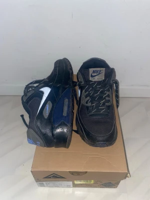 Nike Air Max svarta sneakers - Svarta Nike Air Max 90 sneakers med blå och grå detaljer samt Nike logga på sidan. Ovandelen är i mesh och syntetmaterial, med vadderad plös och häl. Sulan har synlig Air Max-dämpning och reflekterande detaljer på snörningen. Storlek 39