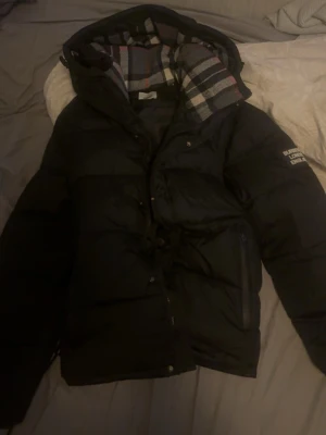 Svart pufferjacka från Burberry - Svart pufferjacka från Burberry med klassiskt rutigt foder i huvan och logga på ärmen. Jackan har dragkedja, tryckknappar och stora fickor framtill. Perfekt för kalla vinterdagar och ger en stilren look.