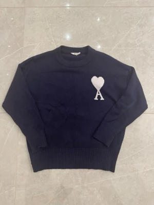 Mörkblå stickad tröja från Ami Paris - Mörkblå stickad tröja från Ami Paris med rund halsringning och ribbade muddar. På bröstet finns ett vitt hjärta med bokstaven A, vilket är signaturdetaljen från märket. Tröjan har en avslappnad passform och är perfekt för lager på lager.