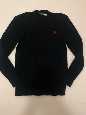 Svart kabelstickad tröja Polo Ralph Lauren - Kabelstickad svart tröja från Polo Ralph Lauren med den klassiska röda loggan broderad på bröstet. Tröjan har rund halsringning och långa ärmar. Perfekt för dig som gillar stilrena och tidlösa plagg.