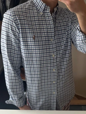Rutig skjorta från Polo Ralph Lauren - Klassisk långärmad skjorta från Polo Ralph Lauren i vitt och blått rutigt mönster. Skjortan har button-down krage, knappar framtill och broderad logga på bröstet. Perfekt för en stilren och avslappnad look. Lite missfärgad i kragen och armhålorna, och några fläckar på armen som man ser på en av bilderna därav priset