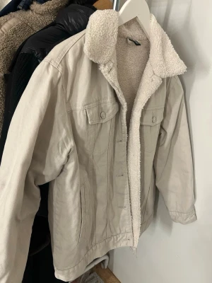 Beige fodrad jeansjacka från H&M - Snygg beige jeansjacka från H&M med mjukt teddyfoder och krage. Klassisk modell med två bröstfickor med lock och knappar samt två sidofickor. Perfekt för dig som vill ha en stilren och varm jacka i jeansmaterial.