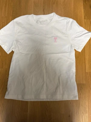 Vit t-shirt med rosa hjärtdetalj Ami - Vit t-shirt från Ami med en broderad rosa hjärtdetalj och bokstaven A på bröstet. Klassisk passform med rund hals och korta ärmar. Tillverkad i mjuk bomull som känns skön mot huden.