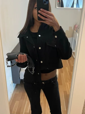 Svart croppad jacka från Zara - Säljer en svart croppad jacka från Zara med silverfärgade knappar och två stora bröstfickor. Jackan har en klassisk krage och är långärmad, perfekt för att ge en cool och trendig look. Materialet är ullblandning som känns mjukt och värmande.