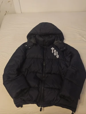 Svart pufferjacka Trapstar London - Säljer en svart pufferjacka från Trapstar London med stor broderad logga och texten 'London Shooters' på ryggen. Jackan har huva, dragkedja framtill och coola detaljer med vita broderier på bröstet. Perfekt för dig som vill sticka ut i vinter.