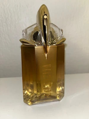 Alien parfym från Mugler guld - Alien parfym från Mugler i en unik, geometrisk flaska med guld- och transparenta detaljer. Flaskan har en futuristisk design med metalliska inslag och tydlig Alien-logga på framsidan. Perfekt för dig som gillar exklusiva och iögonfallande dofter.