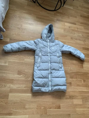 Ljusgrå lång dunjacka med huva - Snygg ljusgrå lång dunjacka med huva och två stora dragkedjeförsedda fickor framtill. Jackan har quiltad design och går ner över låren för extra värme. Perfekt för kalla vinterdagar och har en stilren look. På dragkedjan som du ser pp bilden har det gått bort 2 st tänder där man stänger man kan fixa det för 150kr till 300kr men det går frf att stönga den helt 