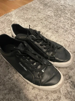 Svarta Calvin Klein sneakers - Snygga svarta sneakers från Calvin Klein med vita sulor och diskret logga på sidan. Skorna har klassisk låg profil, svarta skosnören och är tillverkade i slätt lädermaterial. Använda, dom är för små för mig nu, har växt ut ur dom. Super sköna. 
