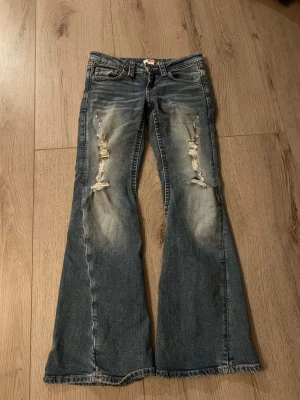 Blå coola bootcut jeans Gina Tricot - Säljer ett par blå bootcut jeans från Gina Tricot med slitningar och hål på låren. Jeansen är i storlek 152🫶Coola o fräcka rosa/röda detaljer på både fram o bak sida!