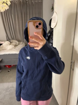 Mörkblå hoodie från Nike - Snygg mörkblå hoodie från Nike med klassisk vit logga broderad på bröstet. Hoodien har en stor huva och en rymlig magficka framtill. Tillverkad i mjukt bomullsmaterial som är skönt att ha på sig. Perfekt för en avslappnad och sportig stil.