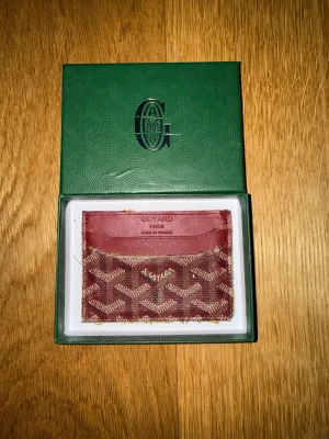 Röd kortfodral från G - Snyggt rött kortfodral från G med klassiskt mönster och logotyp framtill. Fodralet har flera fack för kort och är tillverkat i canvas och läder. Kommer i original grön box med G logga . Perfekt för dig som gillar exklusiva accessoarer.