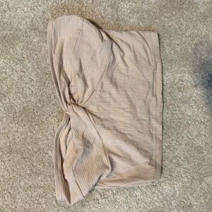 Beige ribbad bandeau tubtopp - Säljer en beige ribbad bandeau tubtopp med knyt framtill. Toppen är axelbandslös och har en snygg twistad detalj som ger en trendig look. Perfekt till sommaren och lätt att matcha med olika outfits.