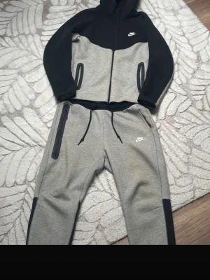 Nike tech fleece set svart/grå - Nike tech fleece set med hoodie och byxor i svart och ljusgrått. Setet har dragkedja på tröjan, fickor med svarta detaljer och justerbar midja på byxorna. Tillverkad i mjukt och stretchigt material, perfekt för träning eller chill.
