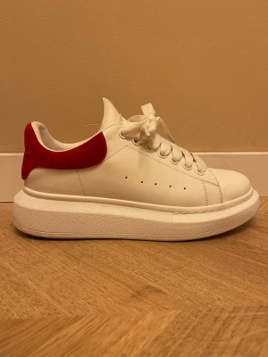 vita sneakers från AM - Stilrena vita sneakers från AMmed chunky sula och röd mockadetalj på hälen. Skon har klassisk snörning, perforerade hål på sidan och logga i silver på hälen. Tillverkad i läder med mocka på bakkappan.