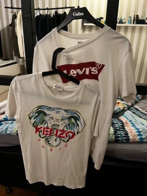 Kenzo och Levi's T-shirt (2 för 1) - Två vita t-shirts, en från Kenzo med färgglatt elefanttryck och röd text, och en från Levi's med klassiskt rött logotryck. Båda har rund hals och korta ärmar. Perfekta för en stilren och trendig look.