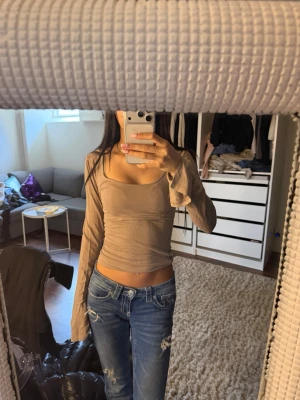 Beige långärmad croppad topp - Trendig beige långärmad topp med croppad passform och smala axelband. Toppen har en figurnära siluett och är tillverkad i ett mjukt material som känns skönt mot huden. Perfekt att styla med jeans för en avslappnad look.