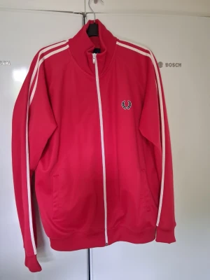 Röd track jacket från Fred Perry - Säljer en röd track jacket från Fred Perry med vita ränder längs ärmarna och vit dragkedja. Jackan har hög krage, två fickor och den klassiska Fred Perry-loggan broderad på bröstet. Perfekt för en sportig och retro stil.