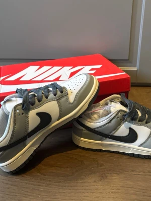 Nike Dunk Low grå och vita sneakers unisex - Nike Dunk Low sneakers i grått och vitt med svart swoosh och svarta skosnören. Klassisk siluett med perforerad tåbox och robust gummisula. Kommer i original röd Nike-låda. Perfekta för dig som gillar streetstyle och sportig look.