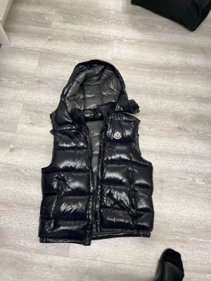 Svart dunväst från Moncler - Snygg svart dunväst från Moncler med glansig finish och klassisk logotyp på bröstet. Västen har två dragkedjeförsedda fickor och quiltad design. Insidan är grå med Moncler-märke och serietidningsdetalj. Perfekt för lager-på-lager och streetstyle.