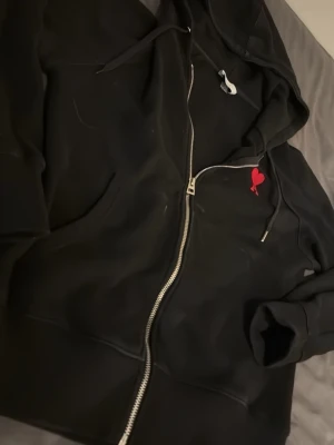 Svart hoodie med röd Ami-dekal - Svart zip-hoodie från Ami med dragkedja framtill, huva med snören och en röd hjärtformad Ami-logga på bröstet. Mjuk bomullskänsla och ribbade muddar vid ärmslut och nederkant. Perfekt för en chill och stilren look. Lite små fläckar, går att få bort.