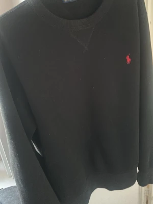 Svart sweatshirt från Polo Ralph Lauren - Svart sweatshirt från Polo Ralph Lauren med klassisk rund halsringning och röd broderad logga på bröstet. Tröjan har ribbade muddar vid ärmslut och nederkant, perfekt för en stilren och avslappnad look.