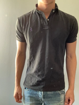 Svart pikétröja från Ralph Lauren - Klassisk svart pikétröja från Ralph Lauren med lila broderad logga på bröstet. Tröjan har korta ärmar, krage och knäppning med två knappar. Tillverkad i bomull för en bekväm och stilren look.