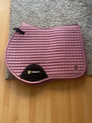 Rosa schabrak från Trinity - Säljer ett rosa schabrak från Trinity med rutmönster och svarta detaljer. Schabraket har en broderad logga och är tillverkat i vadderat tyg, perfekt för ridning och hästutrustning.